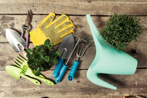 A collection of gardening tools and accessories arranged on a weathered wooden surface, including a pair of yellow gardening gloves, a small trowel and hand fork with blue handles, a pair of pruning shears, a green plant sprayer, and a potted green shrub. The tools are neatly placed with the gloves on top, and the plant sprayer positioned to the right, with the potted shrub nearby. The background highlights a natural outdoor setting suitable for gardening services in Leytonstone, showcasing essential gardening equipment used for planting, cultivating, and maintaining garden beds and outdoor spaces. The scene emphasizes outdoor gardening tasks and the importance of proper tools for garden care, complementing landscaping and gardening service offerings for local residential gardens and outdoor areas.