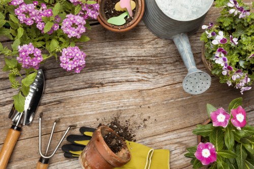 The image depicts a wooden garden table with several gardening tools and supplies arranged on its surface. There are pink and purple flowering plants in small pots positioned around the table, along with a metal watering can and a small terracotta pot filled with soil. A hand rake and gardening gloves are also visible, suggesting recent or ongoing planting activity. The weather appears bright, possibly sunny, with natural outdoor lighting illuminating the scene, which has a natural garden setting that could be part of a landscaped yard in Leytonstone. The arrangement highlights typical gardening steps such as soil preparation and planting, supporting landscaping and outdoor maintenance services provided by Landscapers Leytonstone. The scene underscores a well-maintained outdoor space suitable for garden enhancement or renovation work, fitting within the context of gardening services in the Leytonstone area and postcodes around E11.