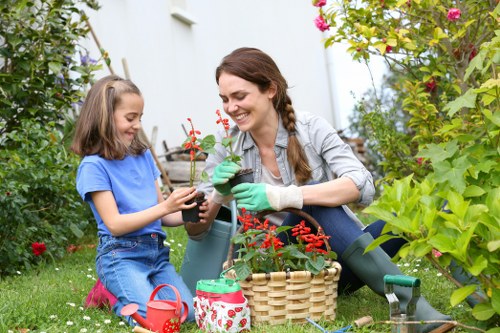 A woman and a young girl are gardening together in a lush, green outdoor space, possibly a backyard garden in Leytonstone. The woman is smiling, wearing green gardening gloves, and holding a small potted plant with red flowers, which she appears to be planting or tending to. The girl, dressed in a blue t-shirt and denim shorts, is also holding a small pot, engaged in the activity, showing interest and enjoyment. Around them, there are various gardening tools and supplies, including a wicker basket, a small watering can, and garden pots. The garden setting features a well-maintained lawn with vibrant green grass, surrounded by flowering shrubs and plants, with some flowers in bloom adding splashes of colour. In the background, tall leafy bushes and a white garden wall create a private and inviting outdoor environment, ideal for family gardening. The scene is illuminated by natural daylight, suggesting a clear or partly cloudy weather, ideal for outdoor gardening activities. This image reflects typical gardening activities that a local Leytonstone gardening service like Landscapers Leytonstone might support through services such as planting, garden maintenance, and lawn care.