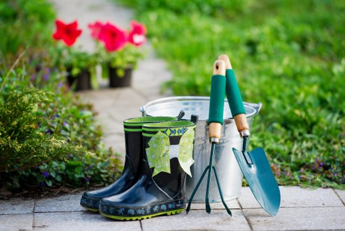 The image depicts a neatly organized outdoor garden scene with a paved pathway bordered by lush green grass and flowering plants. In the foreground, a pair of black gardening rain boots with a decorative blue floral pattern are positioned next to a white bucket. Hanging from the boots are a pair of green gardening gloves. Resting on the ground are a small hand trowel and a larger hand fork, both with wooden handles and metal heads, indicating tools used for planting or soil work. The background reveals a well-maintained garden area with vibrant pink and purple flowers grown in black containers, and a path leading into a green garden environment. The overall setting suggests a prepared outside space suitable for gardening activities, with natural daylight illuminating the scene on a clear day. This image supports gardening and landscaping services offered by Landscapers Leytonstone, connecting to local outdoor maintenance and garden enhancement work in the Leytonstone area.