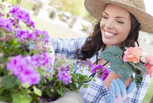 A smiling woman wearing a straw hat and gardening gloves tends to a variety of flowering plants in a well-maintained outdoor garden space, with vibrant purple and pink blooms, lush green foliage, and a neatly arranged paved area visible in the background. The garden features a combination of flower beds and potted plants, with healthy, densely packed grass in the foreground, and the environment appears bright and sunny, indicating clear weather. The scene reflects professional gardening and landscaping activities typical of residential outdoor maintenance services in Leytonstone, as offered by Landscapers Leytonstone, highlighting a tidy, vibrant, and inviting landscaped yard.