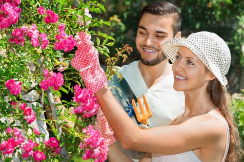A woman and a man are engaged in gardening in a well-maintained outdoor space, with the woman holding a small gardening fork and wearing a polka-dotted sunhat, gloves, and a sleeveless top, while the man stands nearby, smiling and observing. Behind them, there is a vibrant pink-flowered shrub, neatly trimmed hedges, and a lawn area with lush, green grass. The garden is bordered by a combination of paving stones and wooden decking, creating a clean and organized layout. Sunlight filters through the trees, casting natural light on the scene, indicative of a bright, clear day. The environment suggests a residential garden designed for leisure and outdoor maintenance, aligning with professional gardening services such as those offered by Landscapers Leytonstone in the Leytonstone area, near postcode E11, providing insights into typical gardening tasks like pruning, planting, or lawn care.