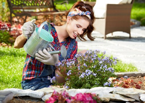 A young woman with a cheerful expression, wearing a plaid shirt, gardening gloves, and a headband, is kneeling in a well-maintained garden in Leytonstone. She is watering a lush, purple-flowered shrub with a metal watering can, surrounded by a variety of plants, including a flower bed with pink blossoms at the front. The garden features a neatly edged lawn with dense green grass, bordered by a low stone edging and a paved pathway. In the background, there is outdoor furniture, including a brown chair on a stone patio, and a wooden fence partially covered by foliage, indicating a private outdoor space. The garden appears to be in good condition, with healthy, vibrant plants and a clean, organized layout, showcasing typical features of residential landscaping services in Leytonstone. Bright natural sunlight illuminates the scene, suggesting a clear, pleasant day in spring or summer, emphasizing the lushness and care given to this outdoor space, likely maintained by local gardening experts such as Landscapers Leytonstone.