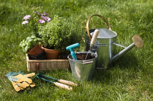 A neatly maintained front garden with a lush green lawn featuring dense, healthy grass. In the foreground, there are garden tools such as small hand rakes and trowels, alongside a pair of gardening gloves, placed on the grass surface. Near the tools, a rectangular wicker basket holds various potted plants and flowering shrubs, including some with purple blossoms. To the right, a metallic watering can with a curved handle and a long spout rests on the grass, suggesting ongoing garden maintenance activities. The scene is outdoors, illuminated by natural daylight, with a well-kept garden layout that includes a mix of grassy areas, potted greenery, and essential gardening tools, likely part of services offered by Landscapers Leytonstone in the local E11 postal district, creating an inviting and functional outdoor space.