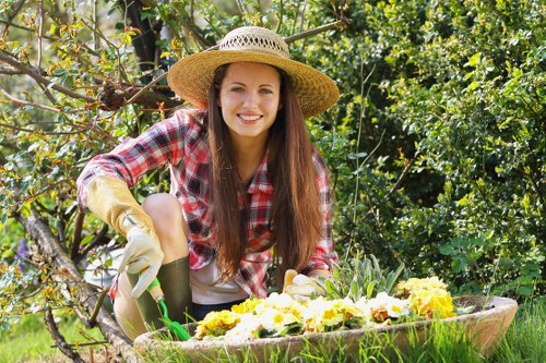 A young woman with long brown hair, wearing a wide-brimmed straw hat, a red and white checkered shirt, and gardening gloves, is kneeling in a well-maintained garden. She is smiling while tending to a flower bed that contains a variety of blooming yellow and white flowers. Behind her, there are lush green shrubs, small trees, and a dense hedge line, indicating a landscaped outdoor space typical of residential gardens in Leytonstone. The garden features a neatly kept grass lawn in the foreground, with a wooden border separating it from the flower beds. Sunlight filters through the foliage, suggesting a bright, clear day, and the environment appears to be in good condition, reflecting professional gardening work, possibly by Landscapers Leytonstone. The scene highlights a healthy, vibrant outdoor area that shows care and maintenance, suitable for lawn care, planting, and general garden landscaping services in the local area.