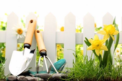 The image showcases a small, well-maintained front garden area with a white picket fence in the background. In the foreground, there is a patch of vibrant green grass and a section of dark soil, bordered by a low, curved edge. To the right, a cluster of bright yellow daffodils with long green leaves add natural colour contrast. On the left side, there are gardening tools resting on the ground, including a small spade with a wooden handle, a hand rake, and a pair of gardening gloves, suggesting recent or ongoing gardening activity. The scene is illuminated by natural daylight, indicating clear weather, and the environment appears tidy and inviting. This setting reflects typical residential landscaping suited for professional gardening and outdoor maintenance services such as lawn care, planting, and general garden upkeep, which a company like landscapersleytonstone.co.uk might offer in Leytonstone or nearby areas.