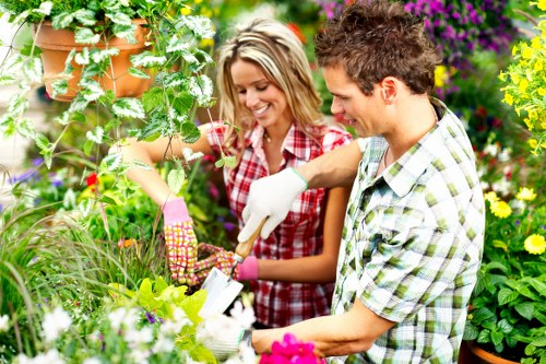 A couple working together in a lush, well-maintained garden with a variety of plants, shrubs, and flowers in full bloom. The woman, wearing pink gardening gloves and a red checked shirt, is holding a gardening tool and tending to a leafy green plant. The man, dressed in a plaid shirt, is assisting with planting or pruning, both smiling as they focus on their outdoor task. The garden features a neatly edged flower bed with vibrant pink, purple, and yellow flowers, alongside dense green foliage and some potted plants hanging from a wooden trellis. The background includes a mix of mature trees and flowering bushes under natural daylight with soft, diffuse sunlight, creating a warm, inviting atmosphere typical of a landscaped backyard in Leytonstone. This scene reflects professional gardening and planting services provided by Landscapers Leytonstone, emphasizing the lush, curated outdoor space, suitable for a garden maintenance or planting project in the local area.