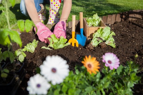 A person wearing pink gardening gloves is working in a garden bed in an outdoor space, planting or tending to young green lettuce or leafy vegetables. The garden area features dark, rich soil and is bordered by a row of small wooden edging planks. In the foreground, there are colorful flowers, including white, orange, and purple blooms, adding visual interest to the garden scene. Behind the person, a set of small garden tools with bright yellow and blue handles are planted into the soil, indicating active gardening work. The background showcases a well-maintained lawn with lush green grass, with dappled sunlight suggesting a bright, sunny day. The overall scene reflects routine garden maintenance or vegetable planting, consistent with professional gardening services provided by Landscapers Leytonstone in the local area, and emphasizes a neat and vibrant outdoor environment suitable for residential or community gardening projects.
