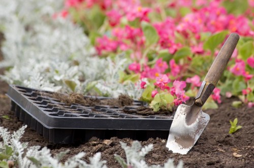 A gardening scene in a landscaped outdoor garden area shows freshly turned soil with a small silver trowel with a wooden handle partially inserted into the earth, and a black seed tray filled with soil resting on the ground. Surrounding the garden bed are clusters of vibrant pink and white flowering plants, possibly begonias or similar ornamental species, creating a colourful backdrop. The garden features neatly maintained soil borders, with visible rich, dark soil indicating recent preparation for planting. The area appears well-lit with natural daylight, suggesting a clear, mild weather day in Leytonstone, contributing to a healthy, inviting outdoor environment. This setup showcases typical elements of gardening and landscaping work, possibly provided by Landscapers Leytonstone, aligning with services such as garden renovation or planting preparation, aimed at enhancing outdoor spaces in the local area.