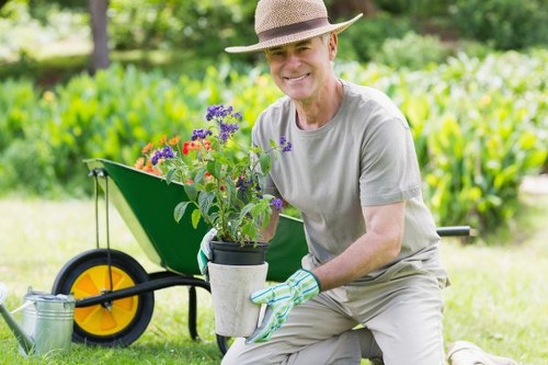 A smiling older man wearing a straw hat, grey t-shirt, and gardening gloves kneeling in a lush outdoor garden area. He is holding a potted plant with vibrant purple and red flowers, appearing to transplant or display it. In the background, there is a well-maintained lawn with dense green hedges and trees, and a green wheelbarrow with a yellow wheel positioned on the grass. The garden features a mix of grass, soil, and densely packed foliage, with natural lighting suggesting a bright, sunny day. The scene reflects a tidy, landscaped outdoor space typical of residential gardens in Leytonstone, suitable for professional gardening and landscaping services by Landscapers Leytonstone.