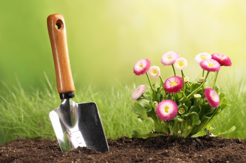 The image depicts a garden scene with a freshly dug bed of rich, dark soil in the foreground, suitable for planting or landscape preparation. On the right side of the image, there is a vibrant cluster of pink and white flowering plants with rounded blooms and green foliage, creating a splash of colour against the natural background. The soil appears well-maintained and free of weeds, suggesting recent gardening activity. In the background, a lush, green lawn extends across the scene, with a blurred natural backdrop indicating a sunny outdoor environment, possibly during late spring or summer. A hand trowel with a wooden handle and metal blade is partially inserted into the soil, signifying active gardening or planting. This scene reflects typical outdoor landscaping work in a residential garden, which could be part of gardening services offered by companies such as Landscapers Leytonstone, providing lawn care, planting, and garden maintenance in the local area near Leytonstone or E11 postcode. The weather appears bright and clear, ideal for gardening activities, reinforcing the image's relevance to outdoor garden maintenance and landscaping services in East London.