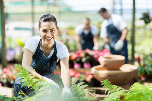 A young woman with dark hair tied back, wearing a white t-shirt and a blue striped apron, is smiling at the camera while working in a well-lit greenhouse or floral nursery environment. She is leaning forward with her hands on a green fern plant, surrounded by various potted flowering plants and foliage. In the background, two other gardeners—one man and one woman—are visible, both wearing aprons and engaged in plant care activities. The setting features glass walls or panels that allow natural sunlight to illuminate the vibrant green leaves and colorful flowers, with shelves and tables holding pots and gardening tools. The outdoor environment behind the greenhouse shows blurred greenery, suggesting a professional garden or nursery in Leytonstone, suitable for landscaping and gardening services offered by Landscapers Leytonstone. The scene emphasizes sustainable plant care and outdoor gardening work, aligning with themes of recycling and sustainability in garden management.